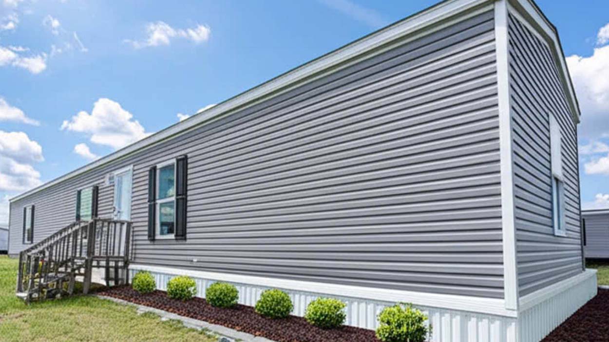 A gray manufactured home with white trim, small front steps, and neatly landscaped bushes along the side.