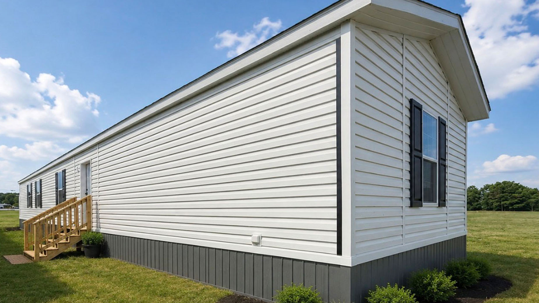 White manufactured home with black shutters and steps, set on grass under a blue sky with scattered clouds, offers comfortable living in a spacious manufactured home.
