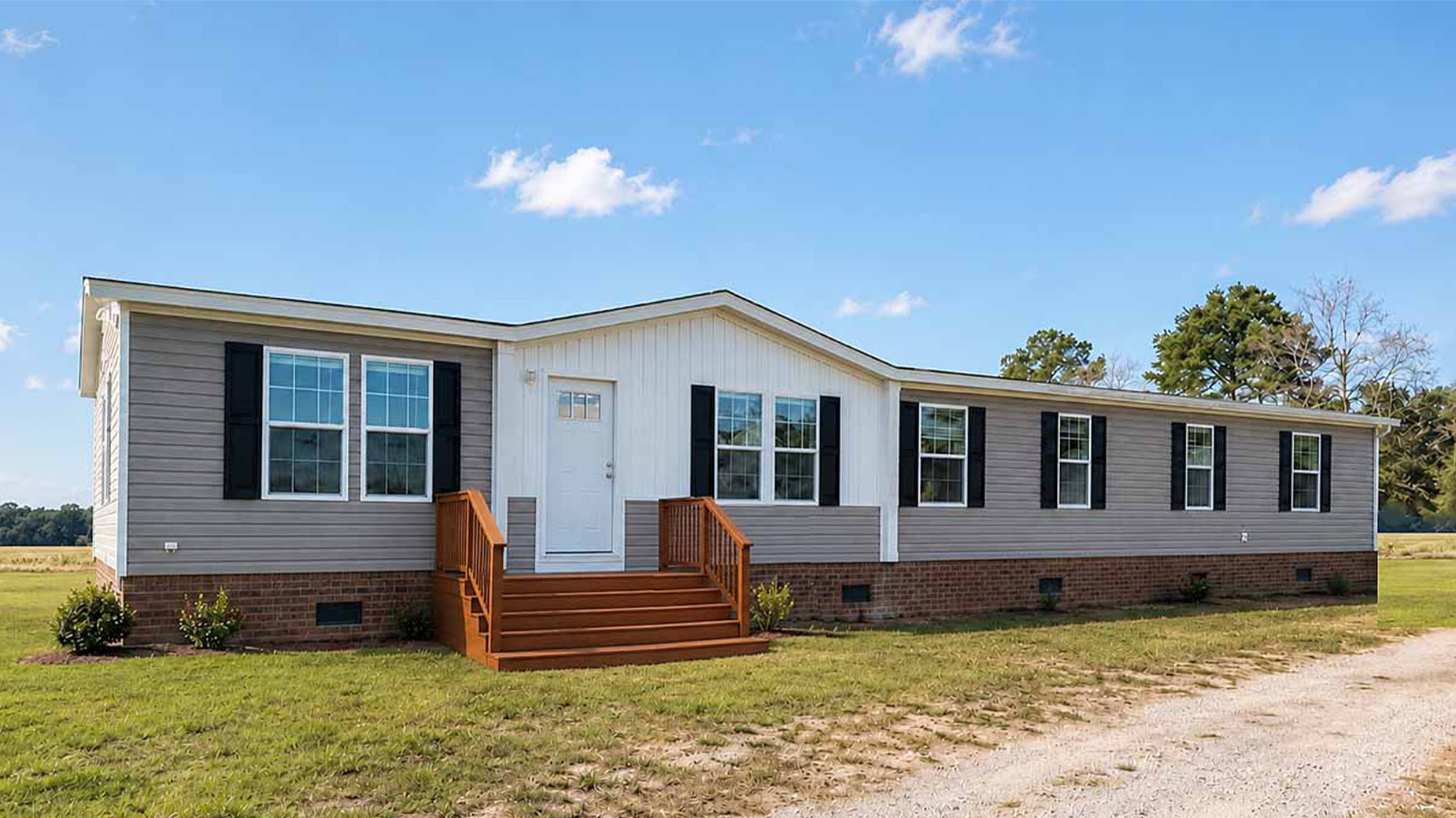 A beige manufactured home with black shutters, brick foundation, and wooden steps sits invitingly on a grassy lot.